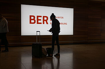 Eine Person steht mit ihrem Gepäck vor einem leuchtenden Flughafenschild am Flughafen Berlin Brandenburg (BER) und blickt auf ihr Smartphone.