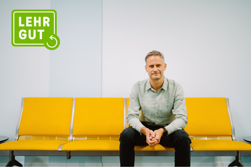 LEHRGUT Interview mit Jörn Scheuren © HTW Berlin/Alexander Rentsch Jörn Scheuren sitzt auf gelber Bank vor weißer Wand mit grünem Logo 'LEHRGUT' in der linken oberen Ecke