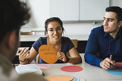 Drei Studierende sitzen mit Stiften in den Händen in einem Seminarraum am Tisch und diskutieren. Auf dem Tisch liegen bunte Pappkarten. Eine Person hält eine mit "critical thinking" beschriftete Karte vor dem Körper.
