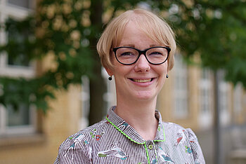 Lächelnde Frau mit Brille und kurz blondem Haar, im Freien vor einem Gebäude mit Bäumen im Hintergrund.
