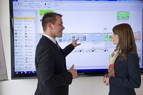 Ein Mann und eine Frau besprechen sich vor einem großen Bildschirm