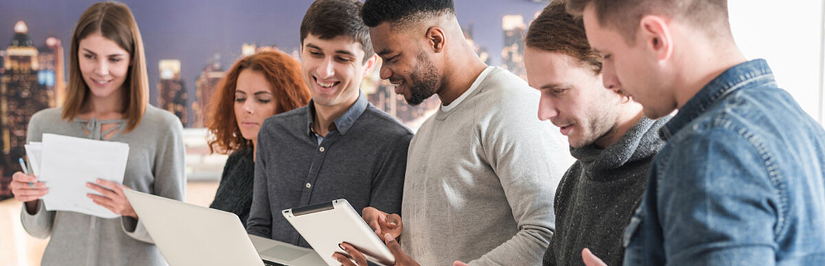 Personen stehen in der Gruppe, halten Laptops und Papier in der Hand und beraten sich gegenseitig