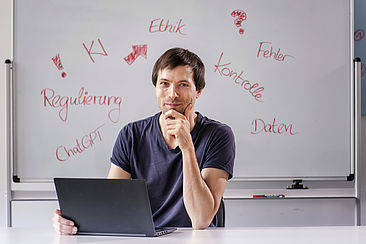 Ricardo Knauer sitzt vor einem Laptop. Hinter ihm steht ein Whiteboard mit folgenden Schlagworten: KI, Ethik, Fehler, Kontrolle, Daten, Regulierung, ChatGPT 