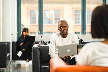 Ein Student arbeitet mit Laptop in einer offenen Lernumgebung, während andere im Hintergrund lernen.