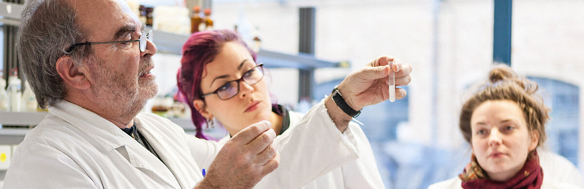 Im Labor: Professor hält ein Reagenzglas und erklärt zwei Studentinnen etwas.