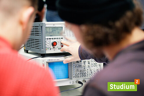 Zwei Studenten vor einem Messgerät, rechts unten das Logo von "Studium hoch 2"