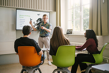 One student at the whiteboard, three students sitting on chairs in front of it, listening