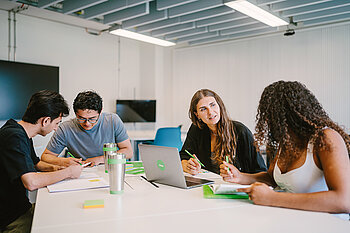 Mehrere Studierende sitzen gemeinsam an einem Tisch und arbeiten mit Unterlagen und Laptops.