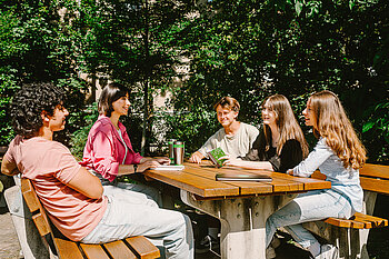 Eine Gruppe Studierender sitzt draußen an einem Tisch und unterhält sich.