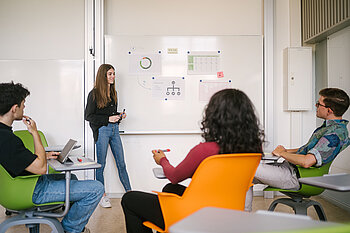 Studierende in einem Stuhlkreis, eine Studentin steht vorn an einem Whiteboard