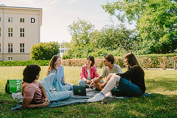 Eine Gruppe Studierender sitzt gemeinsam auf einer Wiese auf dem Campus.
