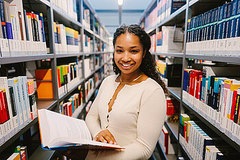Eine Studentin steht zwischen Bücherregalen in der Bibliothek.