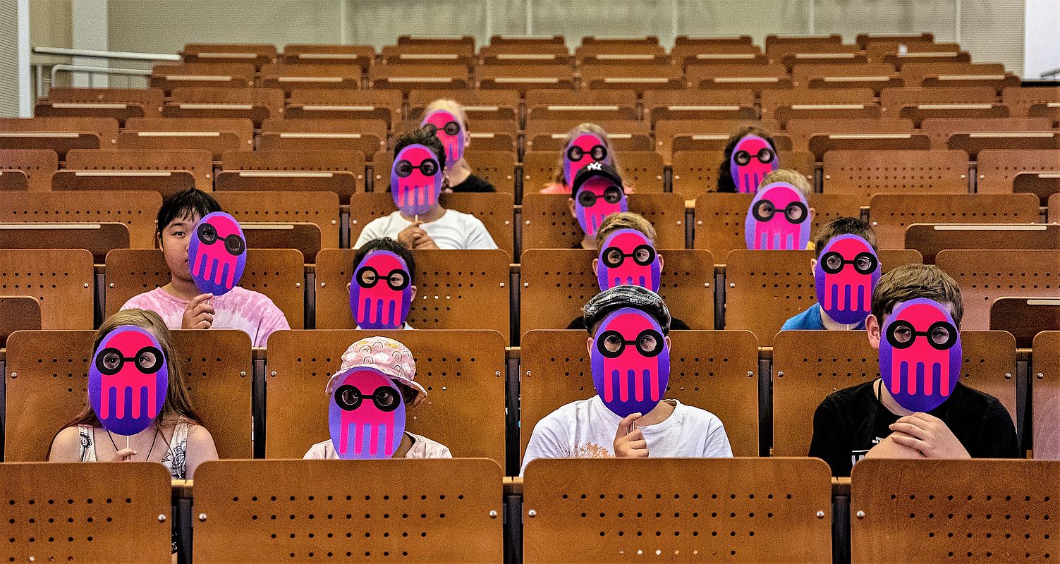 Kinder mit Masken mit dem Maskottchen der Kinderuni im Hörsaal der HTW Berlin © KUL/Dirk Lamprech