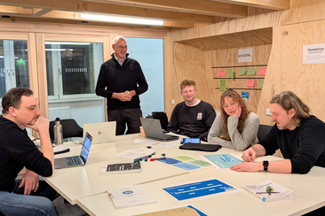 Gruppe von fünf Personen sitzt an einem Tisch und arbeitet gemeinsam mit Laptops, Unterlagen und Moderationskarten, während Prof. Wohlgemuth stehend die Runde anspricht