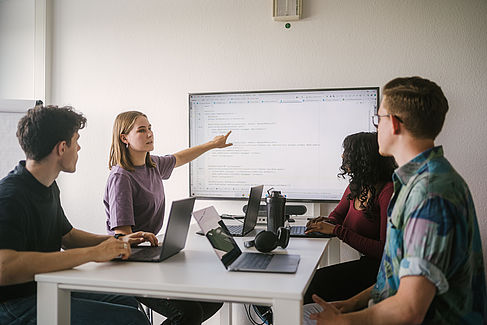 Vier Studierende arbeiten mit Laptops, eine Studentin erklärt etwas und zeigt auf einen großen Monitor