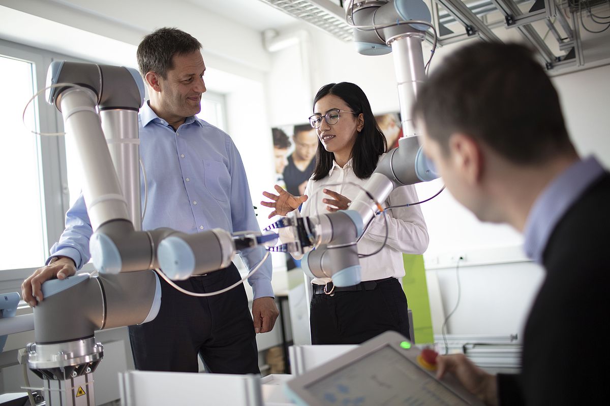 Prof. Schäfer und eine Frau in Business-Kleidung unterhalten sich. Im Vordergrund ist ein Roboterarm zu sehen, der von einem Mitarbeiter gesteuert wird.