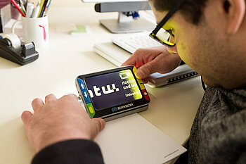 Person hält ein mobiles Bildschirmlesegerät über ein Dokument, das den vergrößerten Schriftzug „tu!“ auf dem Display anzeigt.