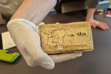 Ein vergilbtes rechteckiges Objekt, beschriftet mit "1899" in einer Hand mit weißem Handschuh